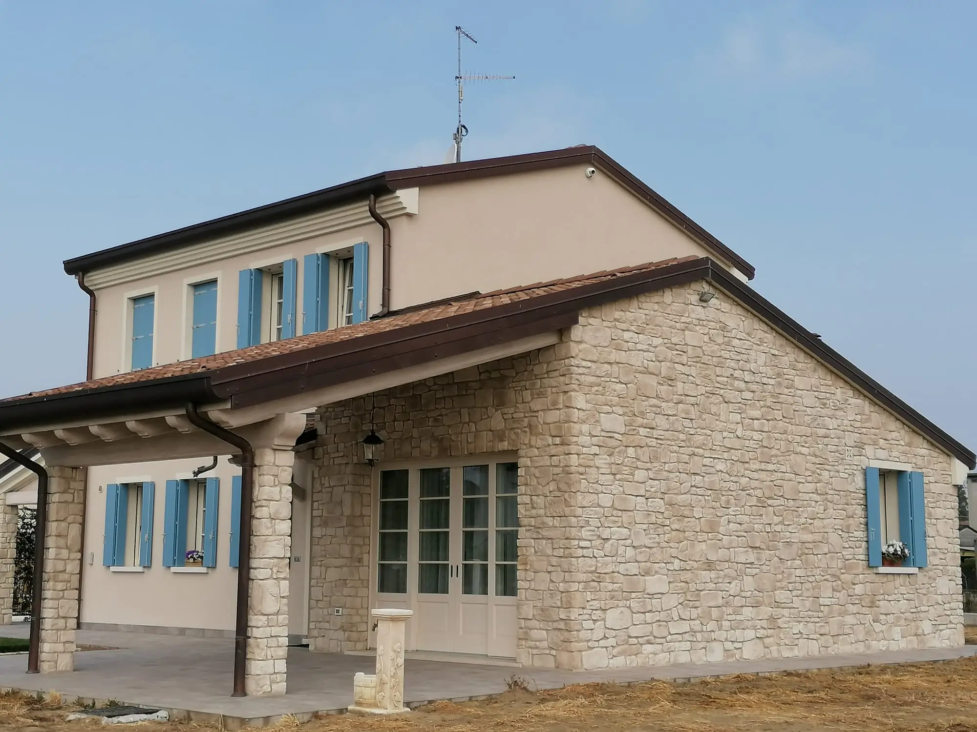Casa rustica italiana in pietra beige con persiane blu e tetto in tegole rosse, vista esterna su terreno arido sotto cielo sereno.