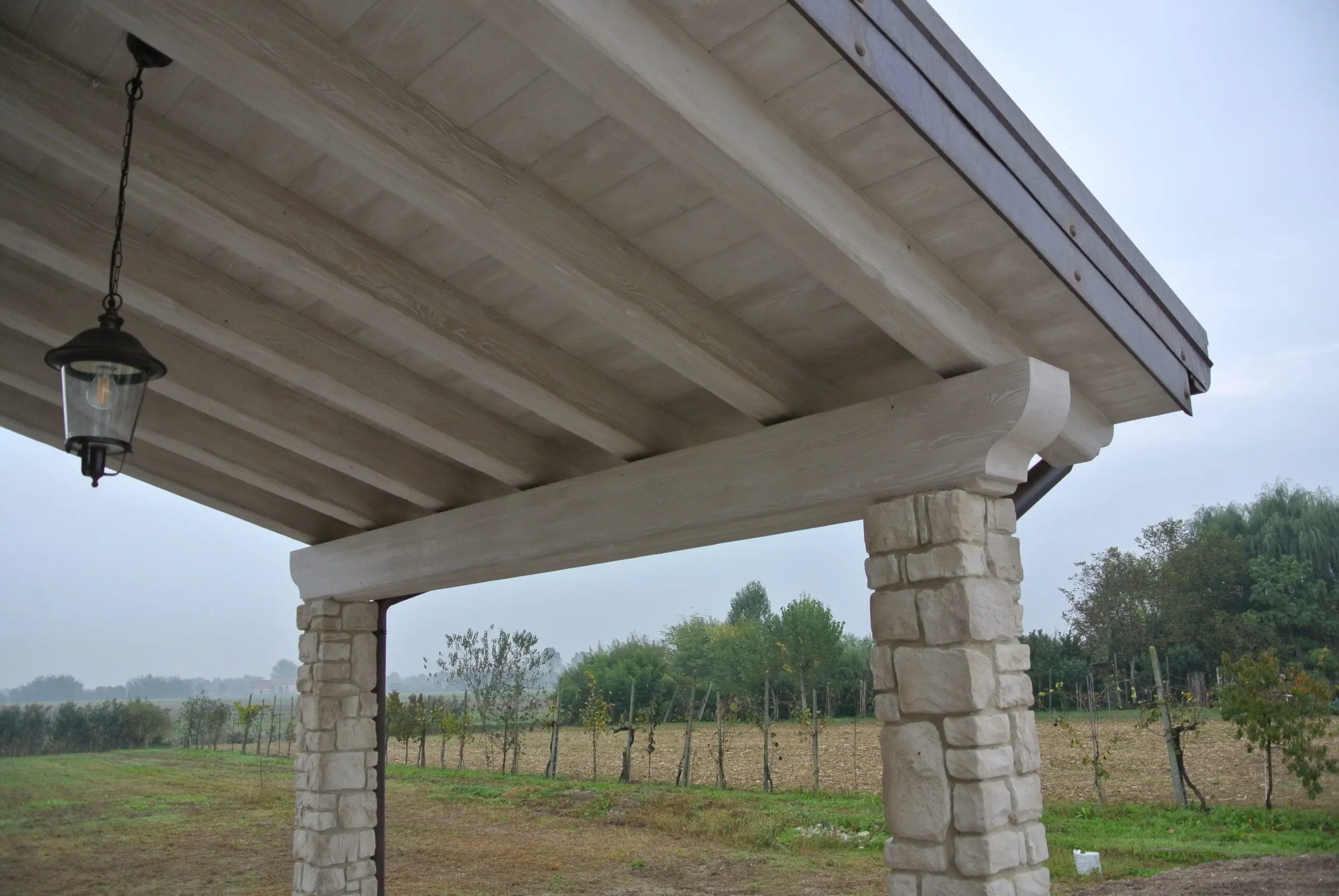 Lampada a sospensione in stile lanterna sotto soffitto portico in legno bianco con colonne in pietra, vista vigneto e campi autunnali.