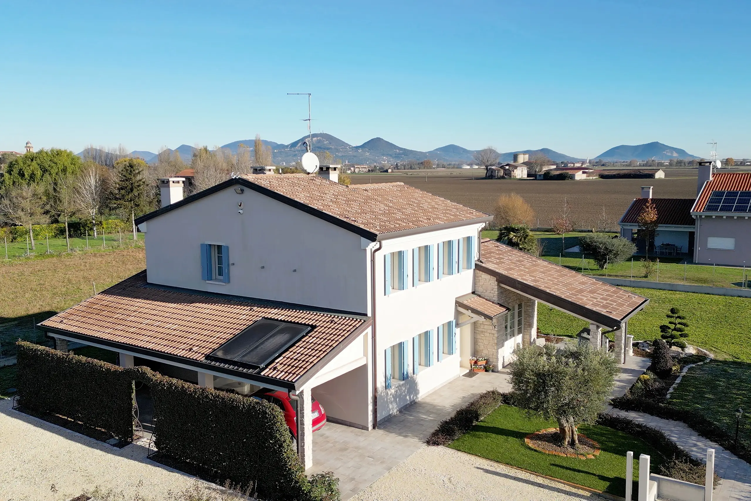 Vista drone bifamiliare ristrutturata progetto ARB: casa bianca intonaco tegole cannucciata portico, orto giardino recinzione, capanno attrezzi, siepi prato, case vicine campi autunnali.