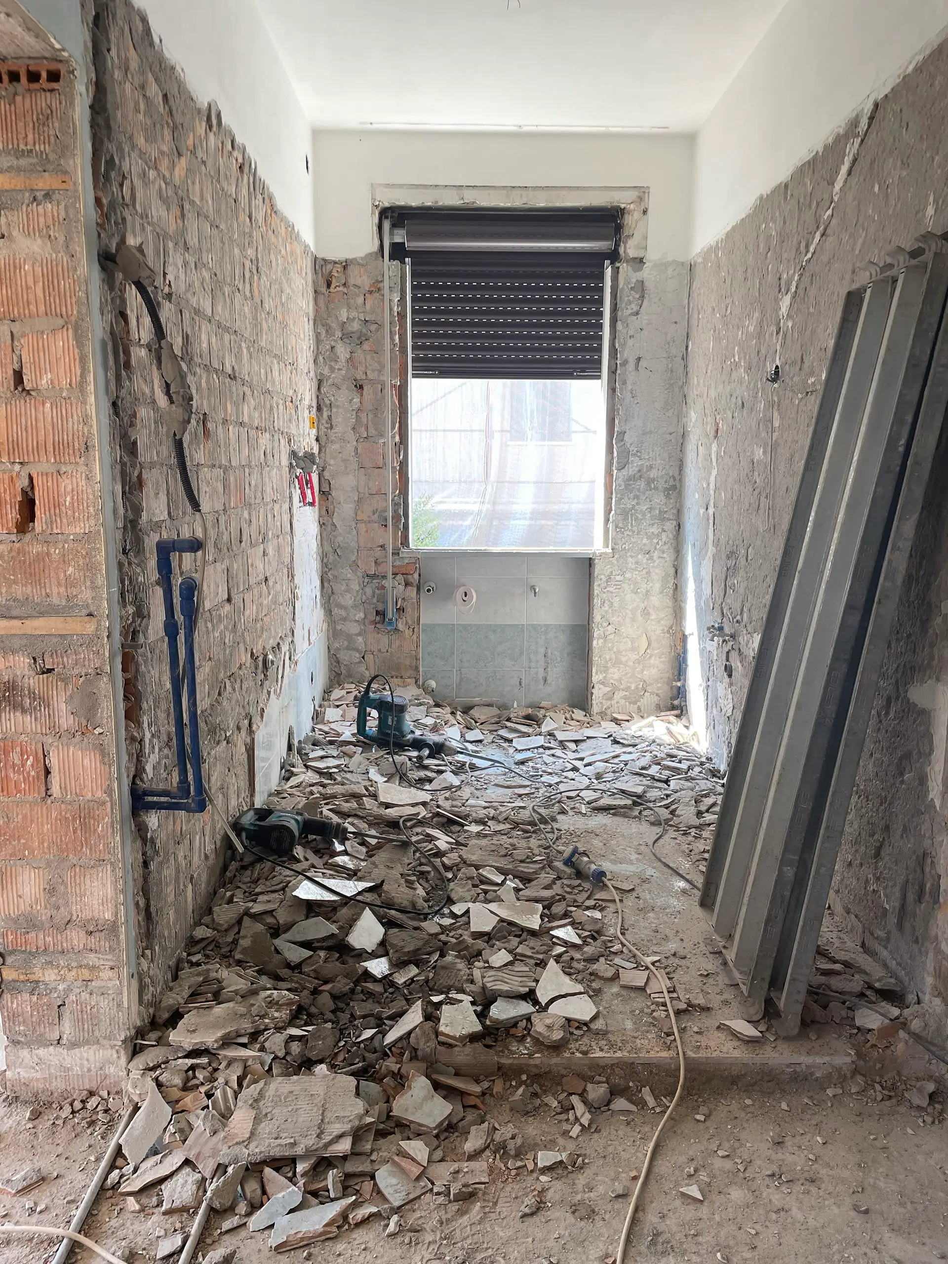Bagno in fase WIP ristrutturazione Casa C.S.: demolizione piastrelle con mattoni a vista, tubature esposte, detriti e impalcature vicino finestra.