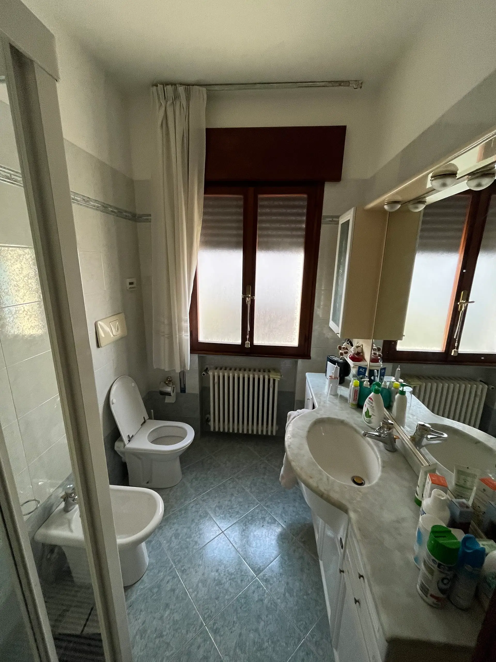 Bagno pre ristrutturazione Casa C.S.: sanitari in ceramica bianca, pavimento azzurro, vasca da bagno e mobile lavabo con prodotti igiene in ambiente datato.