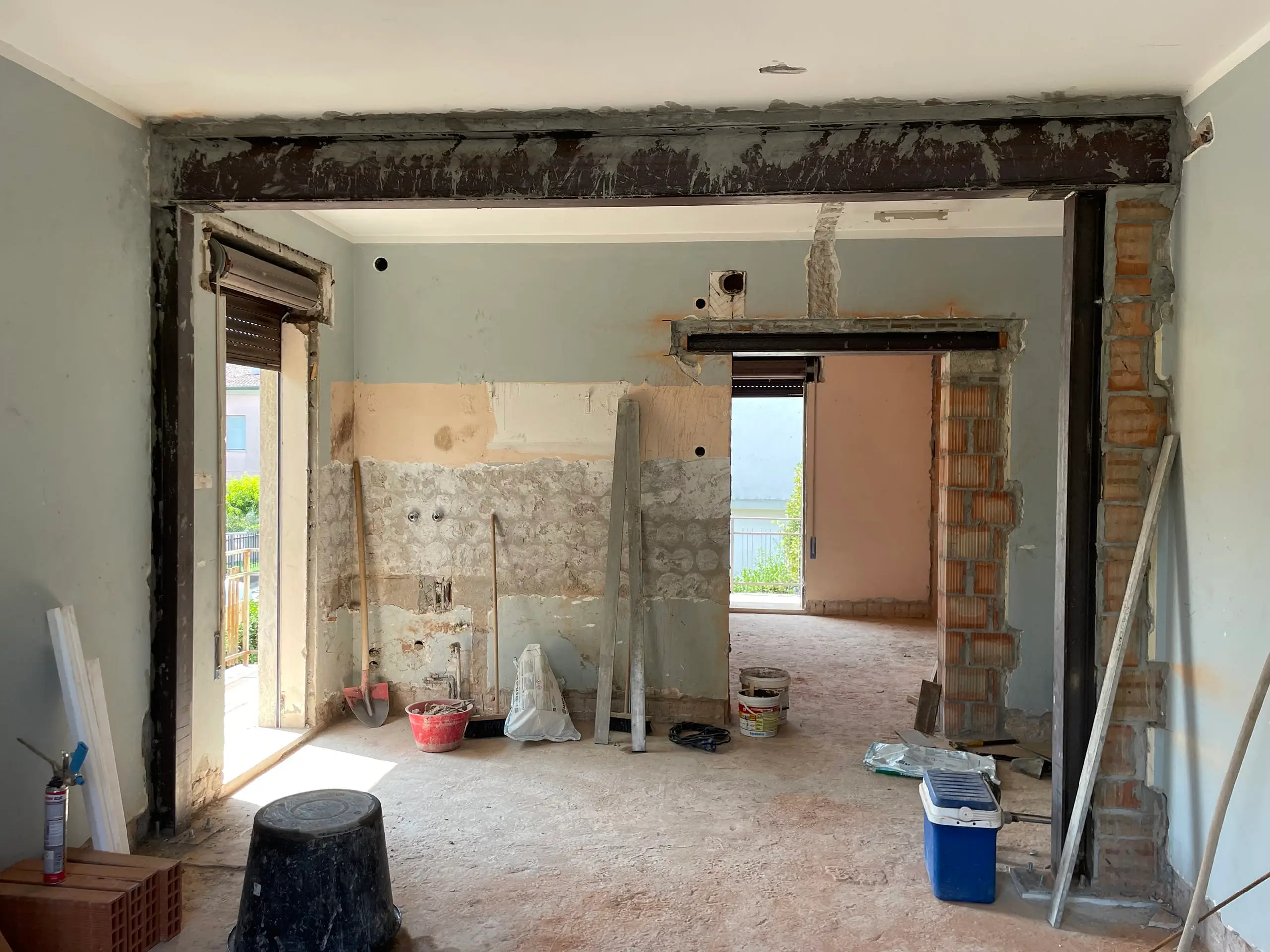 Cantiere in corso WIP zona giorno Casa C.S.: strutture in cemento armato, impalcature, secchi vernice e vista lago attraverso finestre aperte.