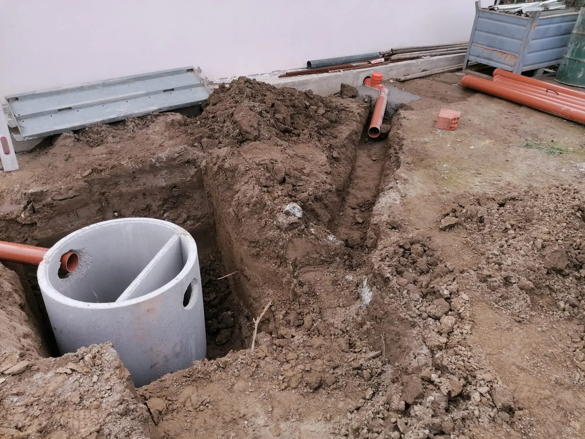 Scavo fossa imhoff depuratore con pozzetto grigio diviso, tubi arancioni fognatura collegati, cumuli terra e materiali edili durante installazione impianto reflui.