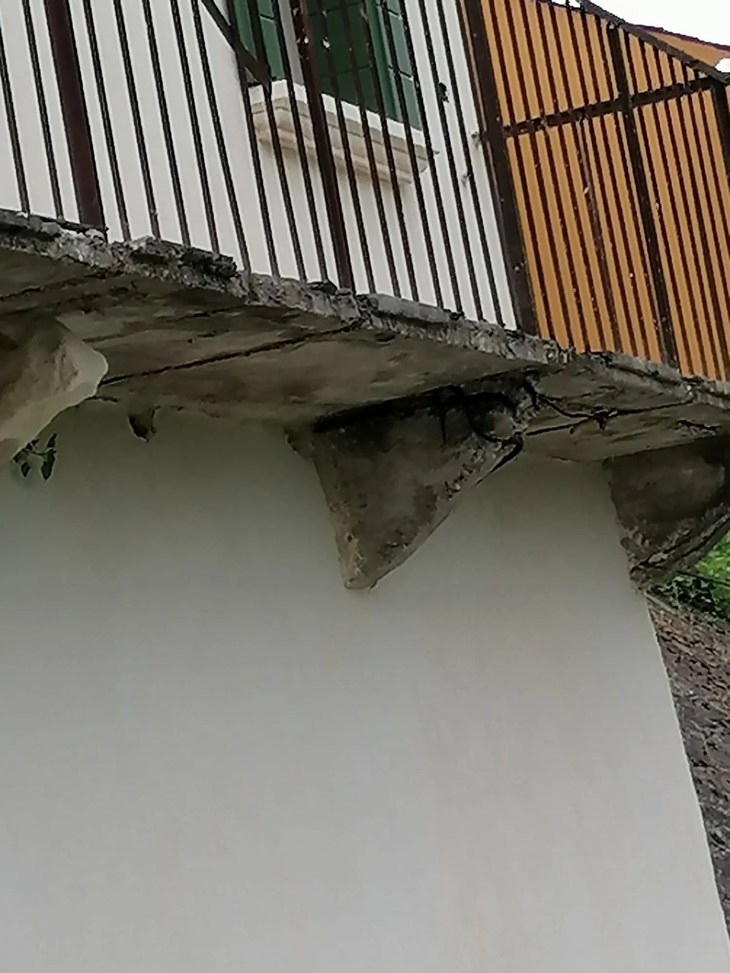 Poggiolo e mensola in calcestruzzo degradati pre-ripristino: ringhiera ferra nera, persiana verde, parete intonacata bianca con crepe e supporto arancione arrugginito.