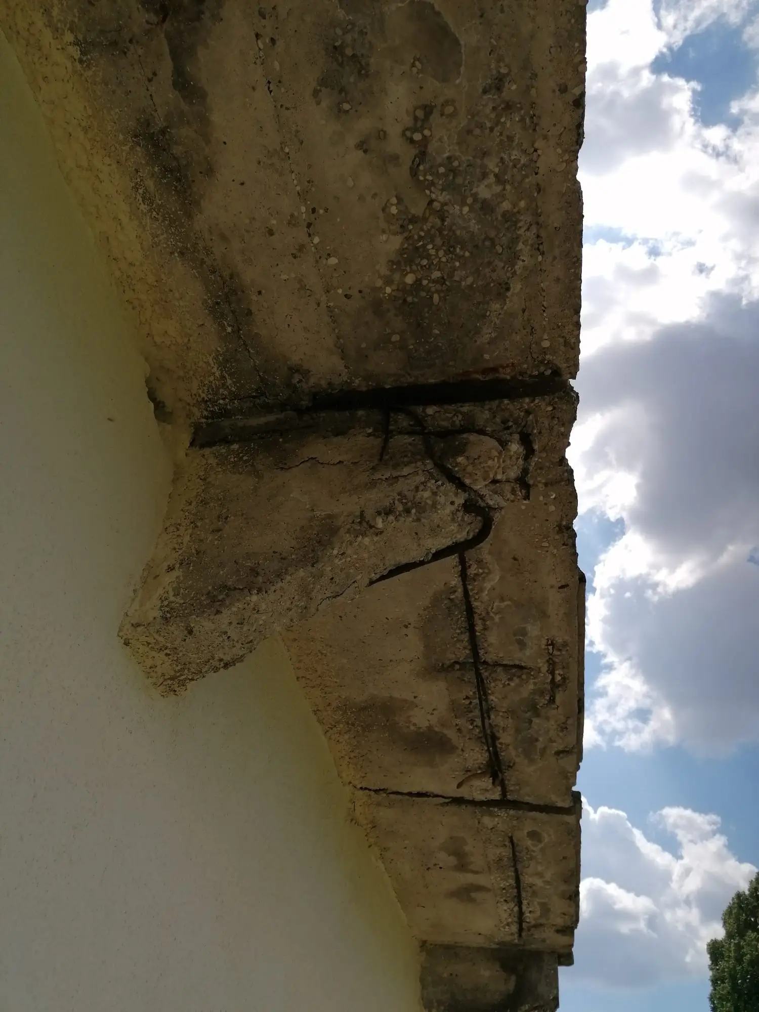 Mensola poggiolo calcestruzzo pre-ripristino con fessure multiple, distacco e ossidazione armatura esposta, parete intonacata bianca sotto cielo nuvoloso.