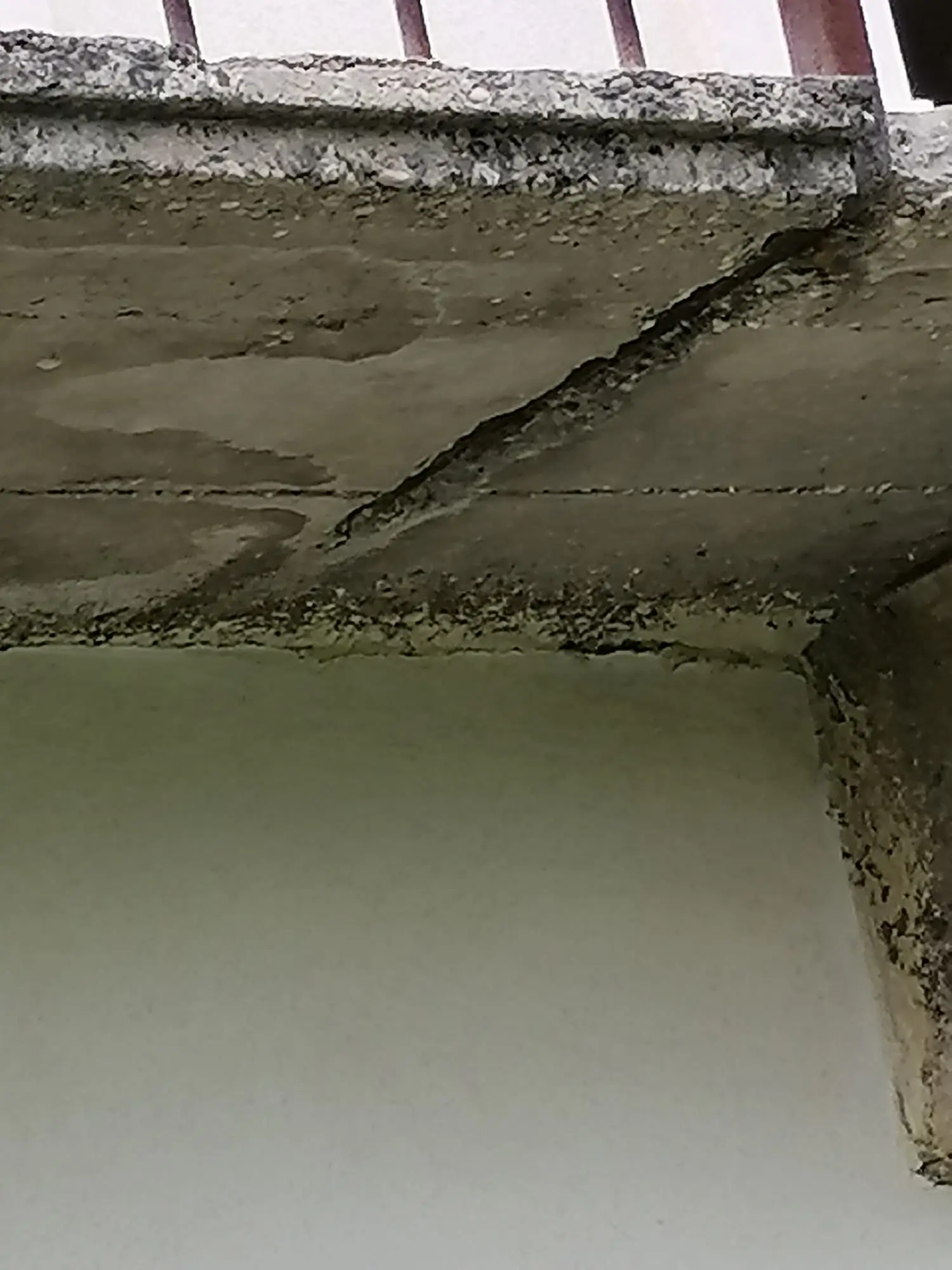 Pavimentazione poggiolo pre-ripristino con crepe longitudinali e distacchi calcestruzzo, ringhiera ferra rossa ossidata, parete laterale chiara cielo nuvoloso.